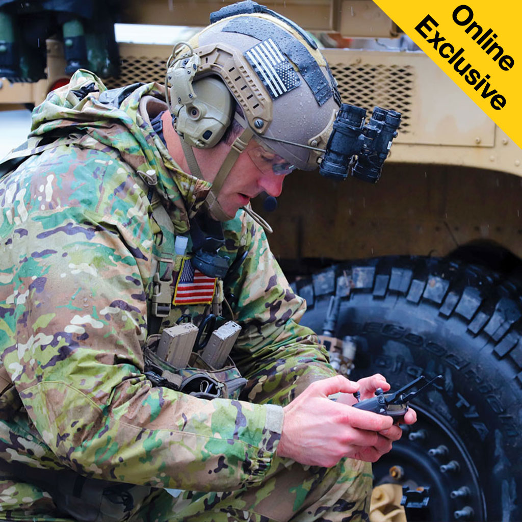A soldier in camouflage gear focuses on a handheld device, with a military vehicle in the background.