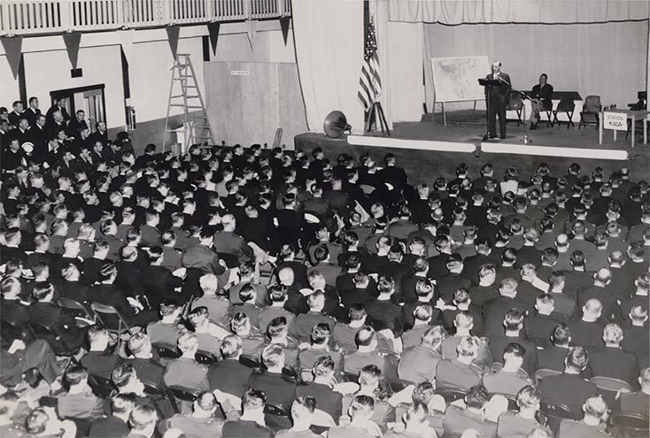 Army and Navy Civil Affairs Staging Area (CASA) officers listen to a civilian speaker on stage with a large map of Asia at an assembly in the spring of 1945 in Presidio of Monterey, California. 