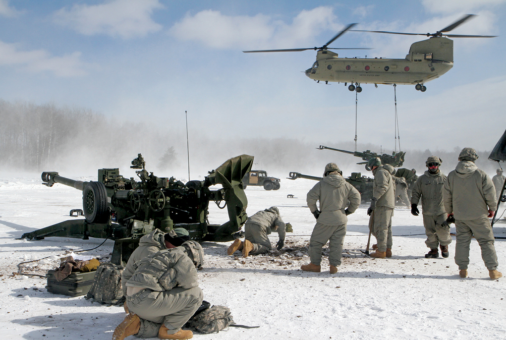 A CH-47 Chinook with a sling loaded M-777 155 mm howitzer flies overhead as soldiers of 1st Battalion, 119th Field Artillery Regiment, use picks to remove inches of ice 1 March 2014 in order to place their howitzer during a live-fire exercise at Camp Grayling Joint Maneuver Training Center, Grayling, Michigan. (Photo by Staff Sgt. Kimberly Derryberry, Michigan National Guard)