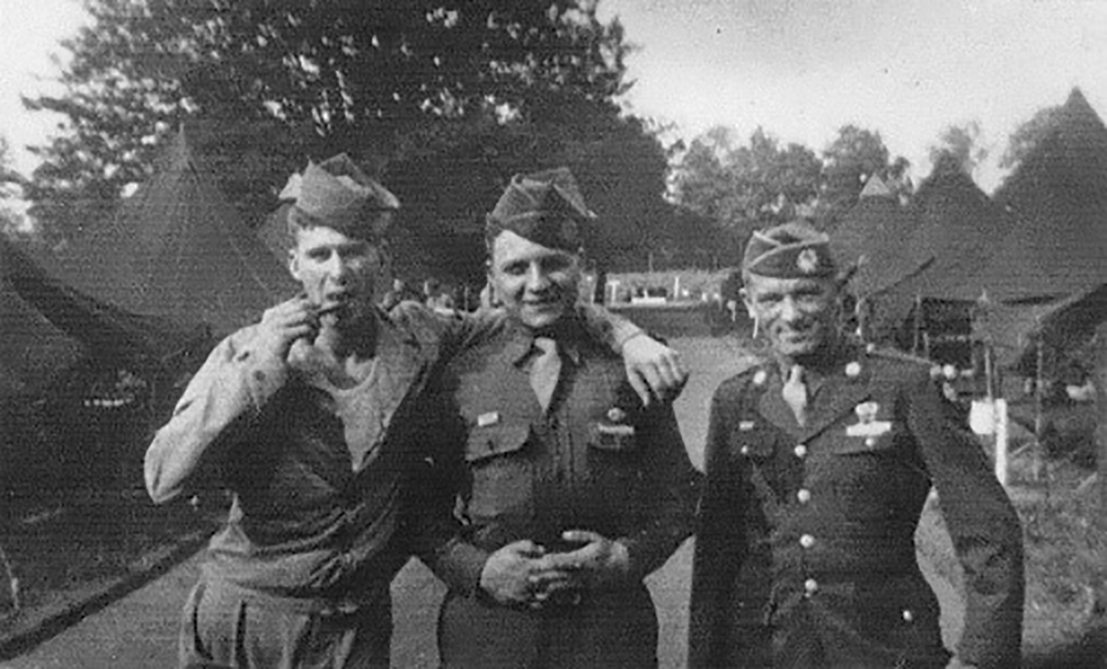 Soldados John E. Atchley (à esquerda), Nicholas J. DePalma e Joseph J. Comer (à direita), paraquedistas da Companhia H, 505o Regimento de Infantaria Paraquedista, no acampamento da unidade, em maio de 1944, várias semanas antes do Dia-D, no Camp Quorn, Leicestershire, Inglaterra. Atchley foi condecorado com o Distinguished Service Cross por suas ações em um ponto de controle em Sainte-Mère-Église, em 07 Jun 44. A citação da condecoração (abaixo) demonstra como Atchley personificou o tipo de iniciativa e a propensão para agir que o treinamento para o comando de missão visa a promover.