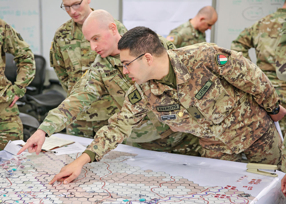 El mayor Stefano Catania del Ejército de Italia (der.) y el mayor Keith Weaver, Ejército de EUA, discuten posibles posiciones para sus tropas mientras el mayor Colin Bair, Ejército de EUA, (al fondo, izq.) observa un juego de Landpower en la Escuela de Comando y Estado Mayor del Ejército de EUA en Fort Leavenworth, estado de Kansas. Landpower, un juego de mesa con hexágonos basado en un mapa, fue probado del 9 a 15 de marzo de 2018 para evaluar su potencial para reemplazar o complementar el simulacro computarizado actual que toma más tiempo y mano de obra para operar. (Foto: Capitán Charlie Dietz, Ejército de EUA)