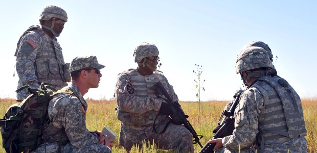 Army military members huddled in group of 5 in a field.