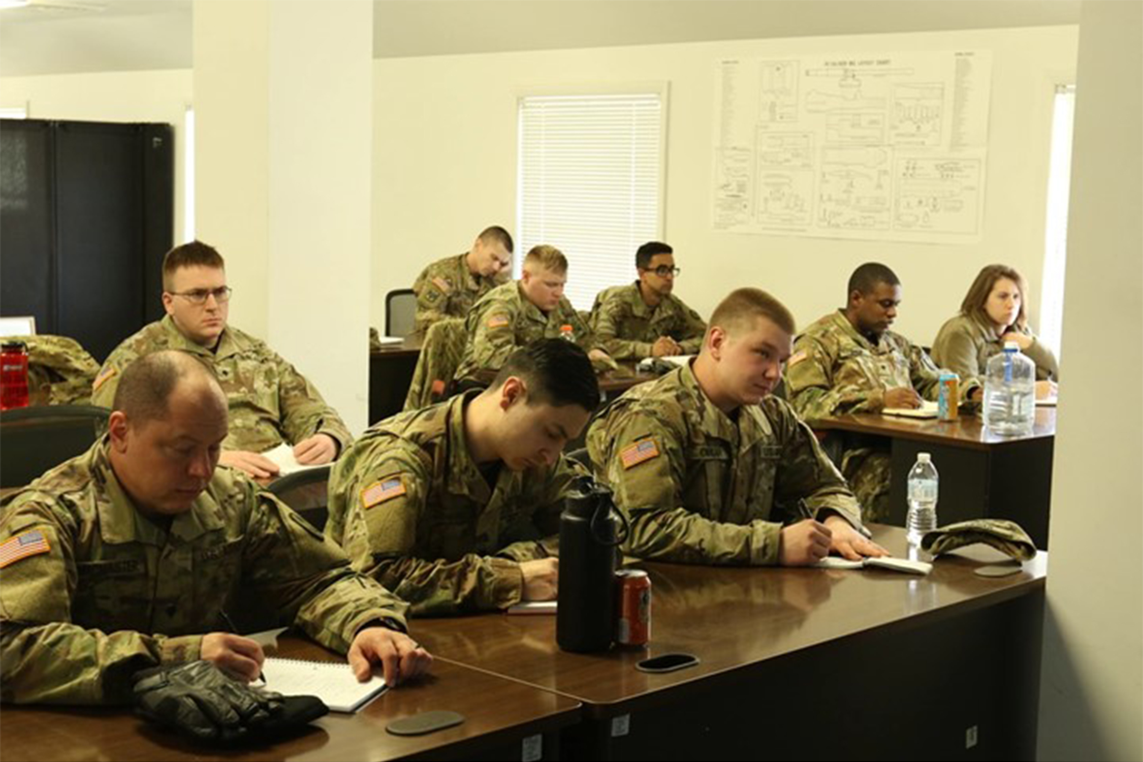 Group of soldiers in uniform attending a training or classroom setting, taking notes.