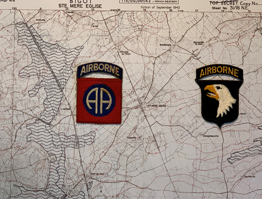 
Two U.S. Airborne Division patches, the 82nd with red and blue 'AA' insignia and the 101st with a black and gold eagle, overlaid on a topographic map of Sainte-Mère-Église, Normandy.