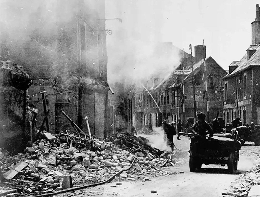 
The image shows a scene of destruction in a town during World War II, with buildings heavily damaged and smoke rising in the background. Soldiers and a military vehicle are visible on a street littered with debris.