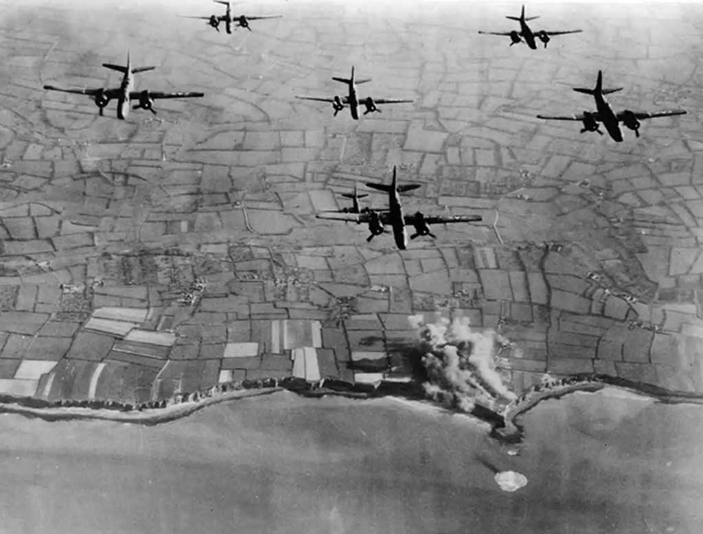 A squadron of World War II bombers flying in formation over a patchwork of fields, with bomb explosions visible below, capturing a moment of aerial warfare.