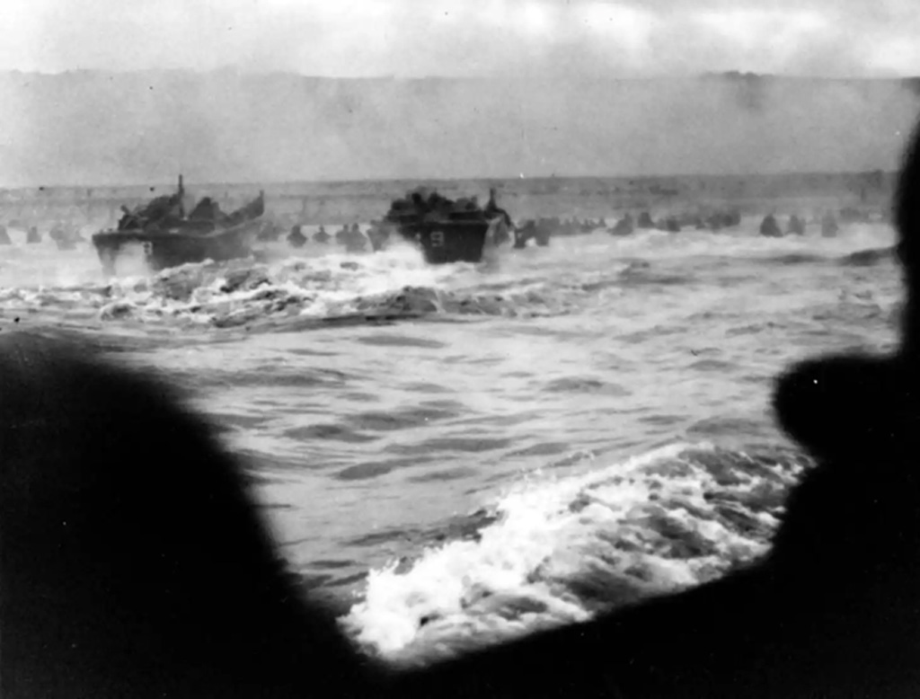 Black and white historical photo of Allied landing craft approaching Normandy Beach with troops inside during World War II, viewed from another craft's ramp.