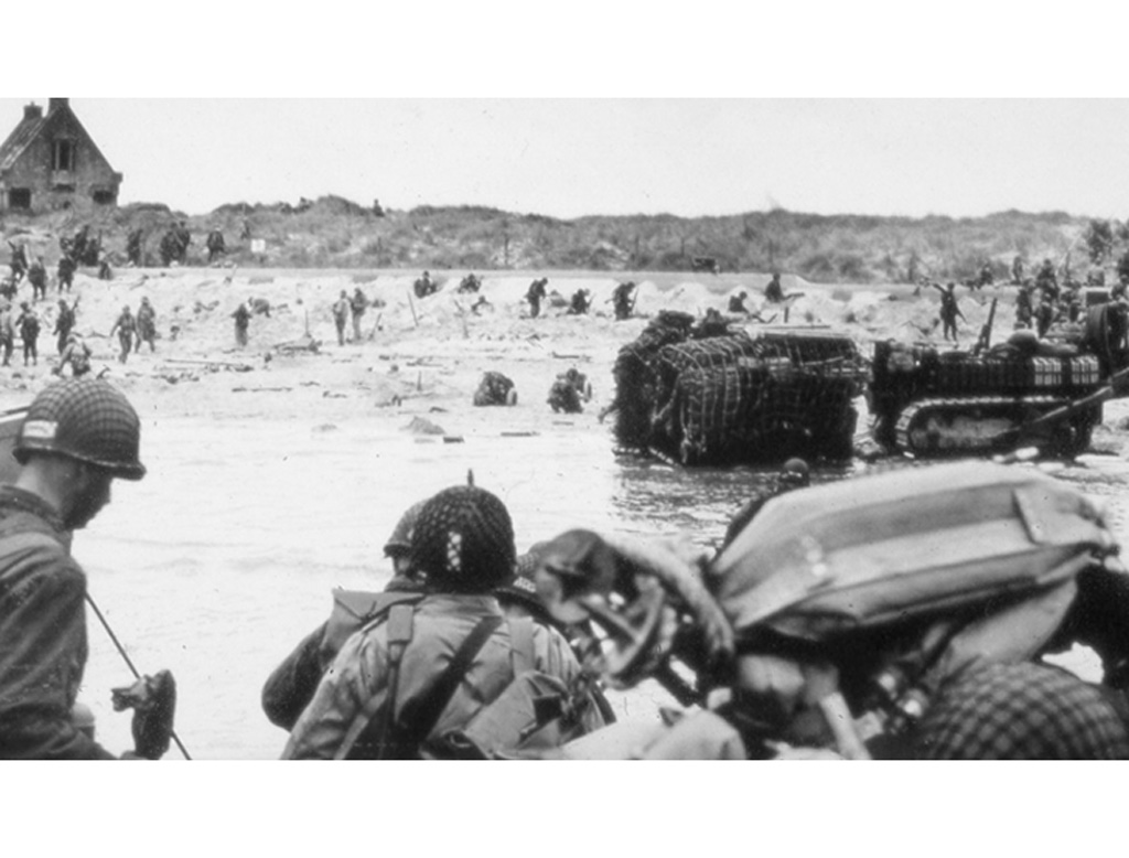 Allied troops landing on a Normandy beach with obstacles, under fire, some seeking cover behind hedgehogs, others moving inland, with a house visible in the background.
