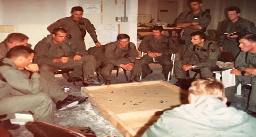 Military personnel gathered around a sand table in a briefing room, discussing strategy or receiving orders, with some taking notes.
