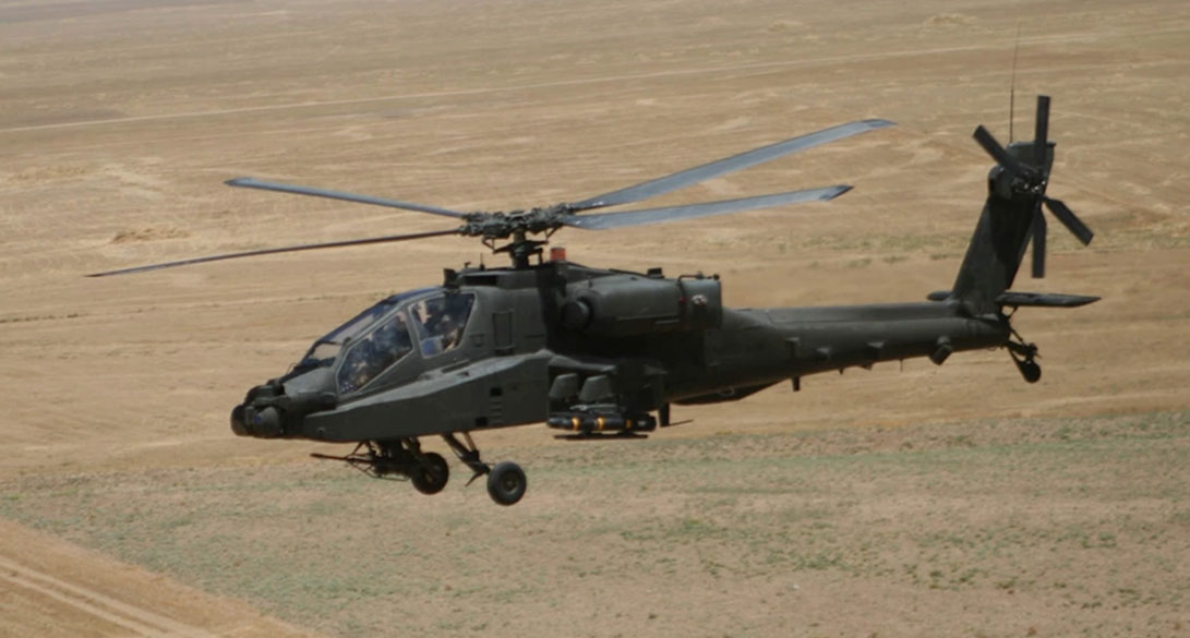 An Apache helicopter flying low over a barren landscape, with its rotors spinning and equipped with armaments.