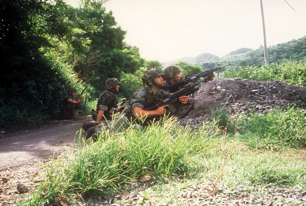 Members of the 82nd Airborne Division take cover behind a gravel embankment while patrolling a road during Operation Urgent Fury