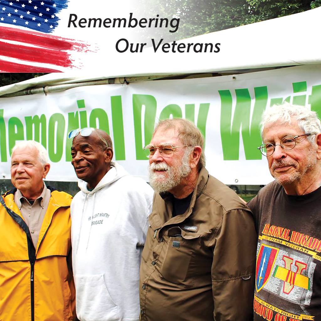 Four veterans standing next to an outdoor tent in casual attire. The banner hanging from the tent reads 'Memorial Day Writers'.