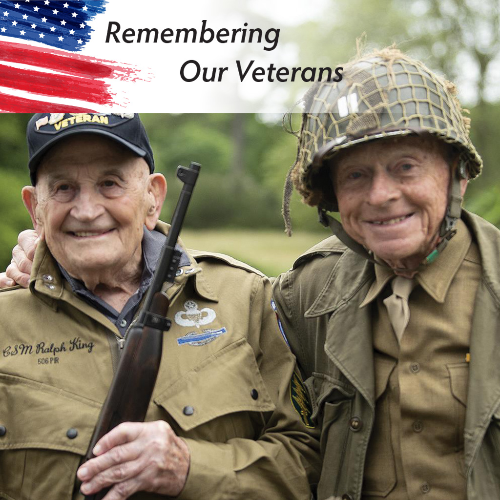 Two smiling elderly veterans in military uniforms, one holding a rifle, with an American flag and 'Remembering Our Veterans' text overhead.