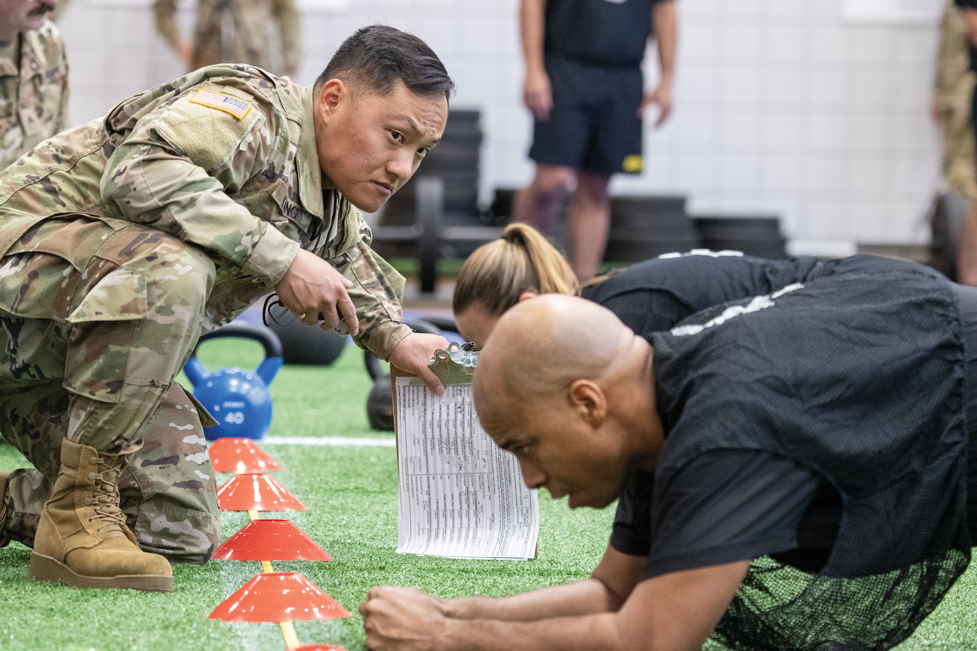 Army fitness assessment showing soldier in combat uniform holding stopwatch and scorecard while supervising participant performing plank exercise on artificial turf. Orange marker cones and kettlebells visible. Other participants and observers in background. Demonstrates Army Combat Fitness Test evaluation procedures.