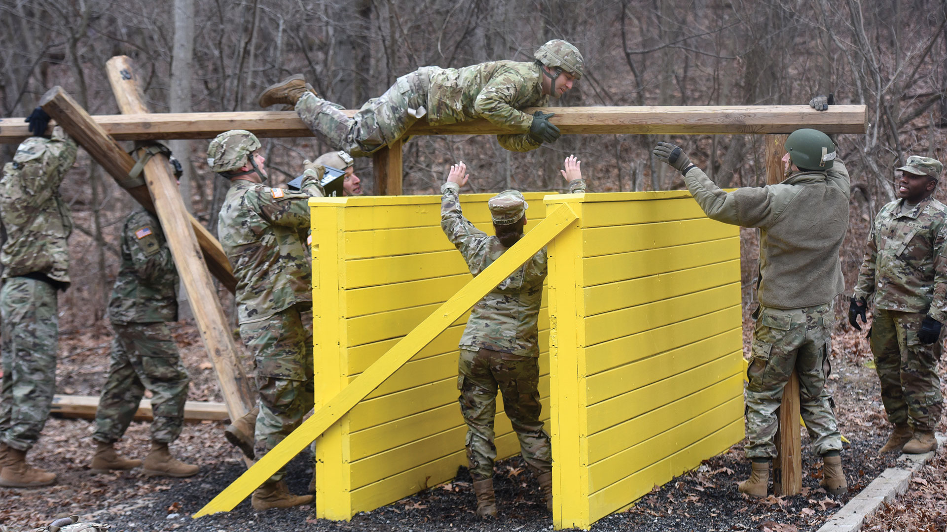 Soldiers completing obstacle course challenge featuring yellow wooden wall. One soldier traverses horizontal beam at top while team members below assist and spot for safety. All wearing multicam uniforms in wooded training area. Demonstrates teamwork and physical fitness training during leadership course.