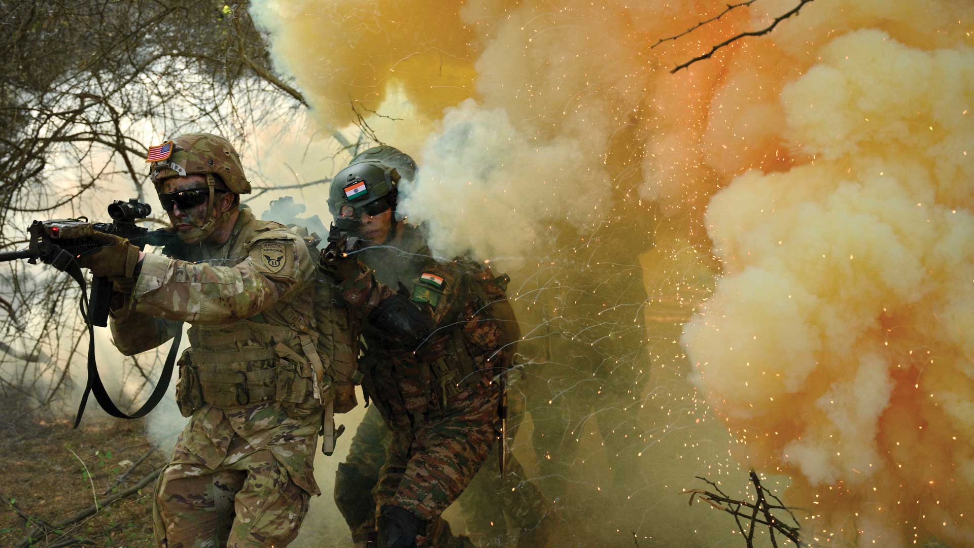 U.S. and Indian soldiers in camouflage gear advance through smoke and sparks in a wooded combat training scenario.