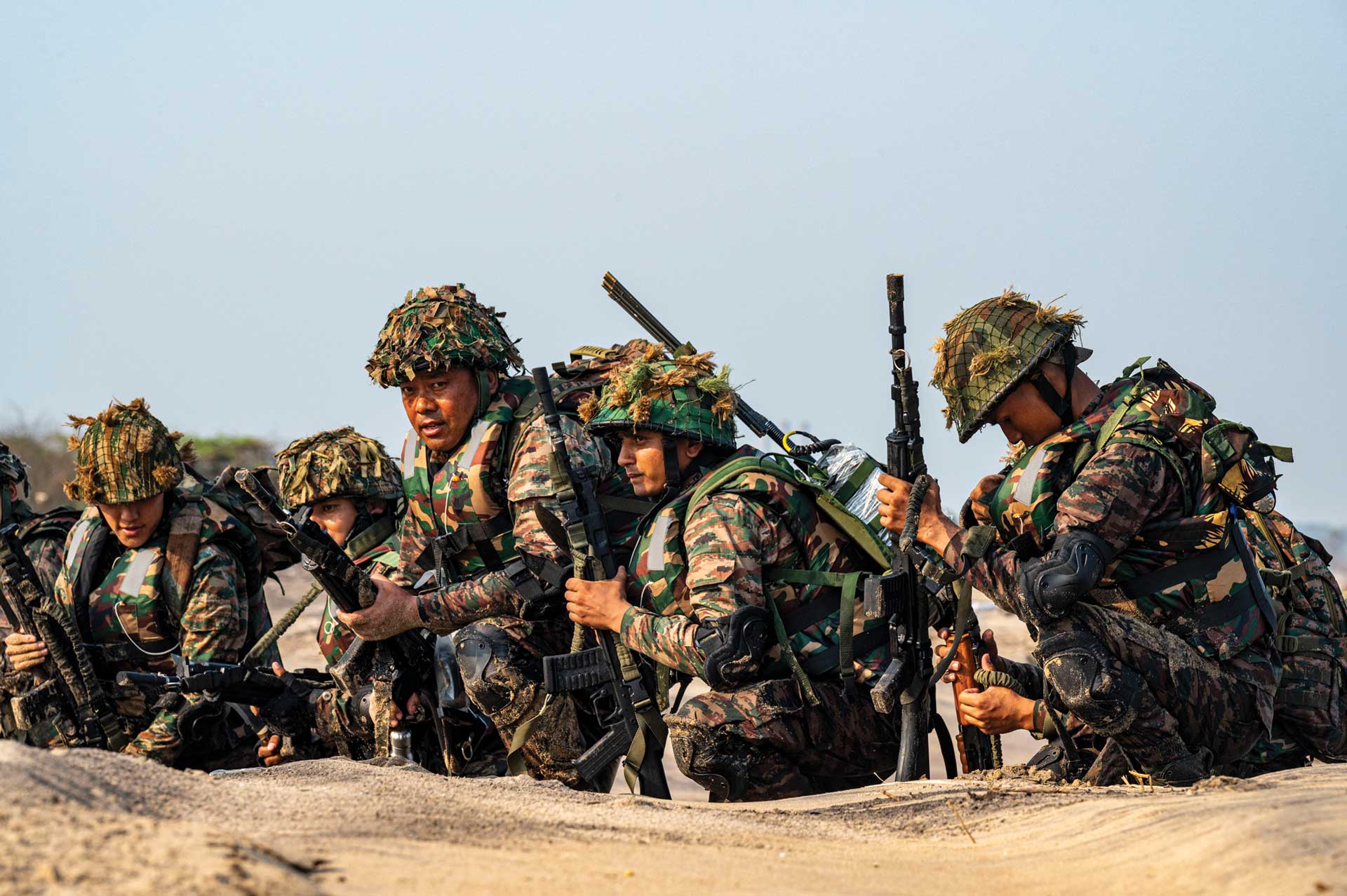 Indian soldiers in camouflage gear crouch in a sandy terrain during a tactical exercise, holding rifles.