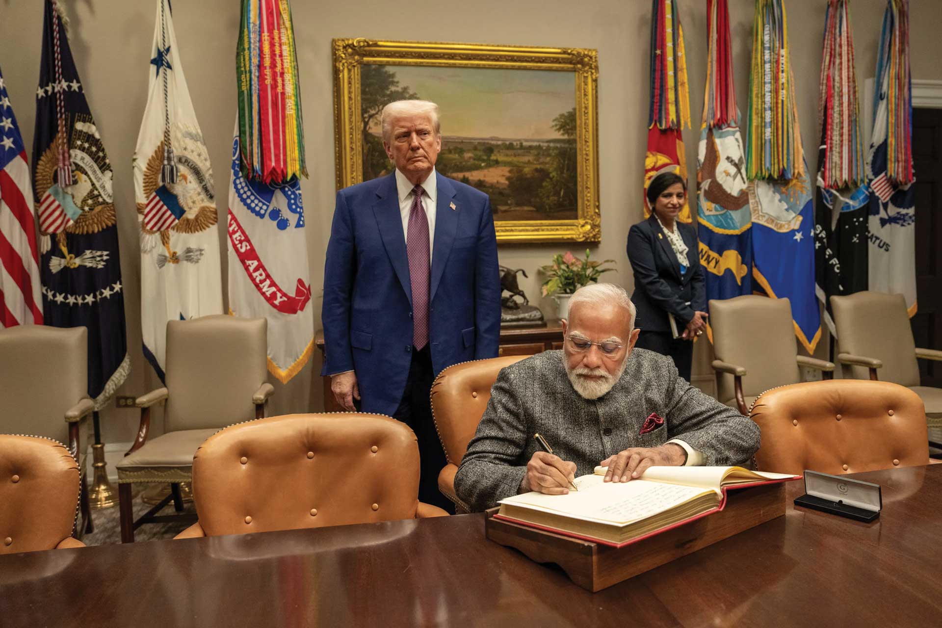 Indian Prime Minister signs a book at a formal table while U.S. President Donald J. Trump stands behind him in a room with military flags.