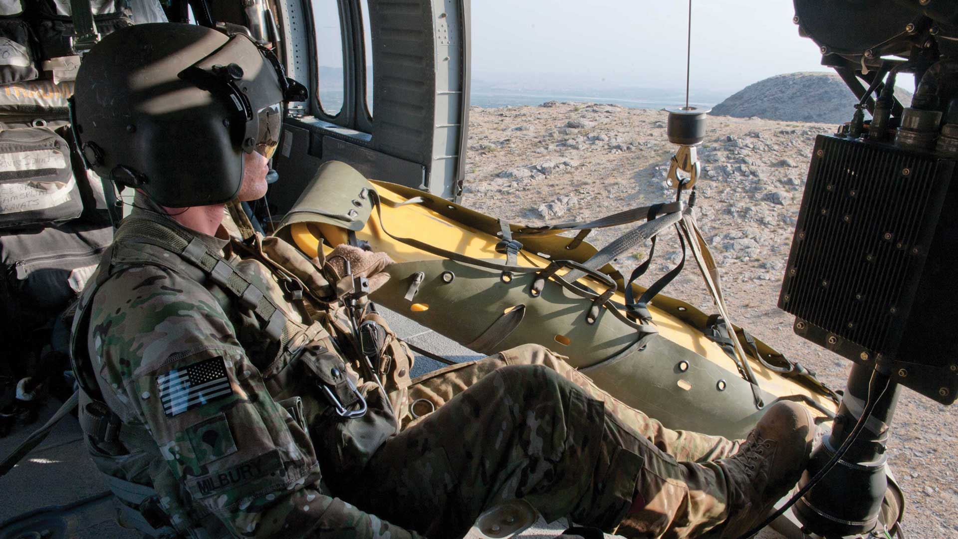 A U.S. Army soldier inside a military helicopter monitors a rescue litter hanging out the side door during a medical evacuation mission over rocky terrain.