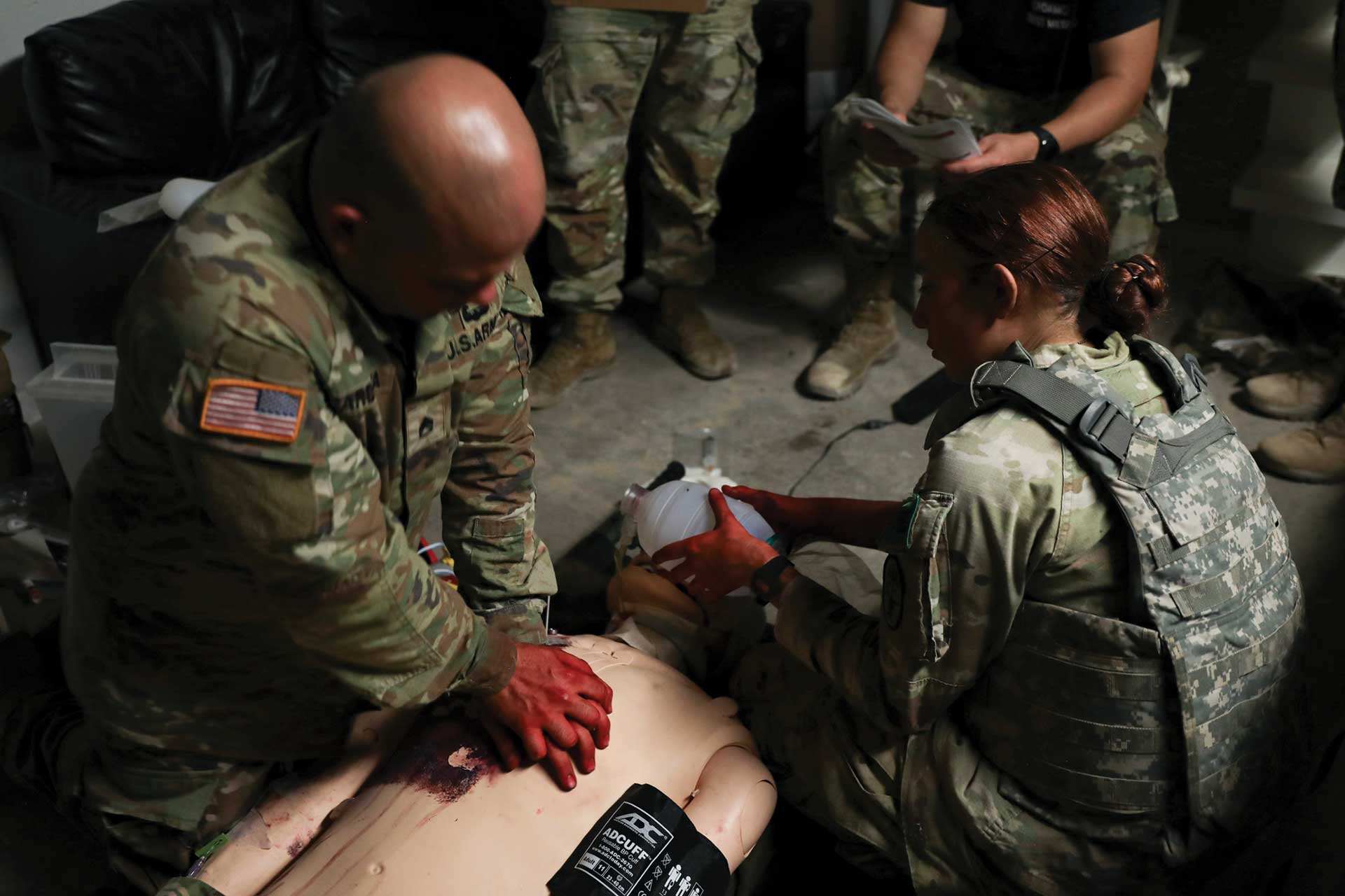 Military personnel perform CPR on a medical training dummy with visible wounds during a simulated combat casualty care exercise indoors.