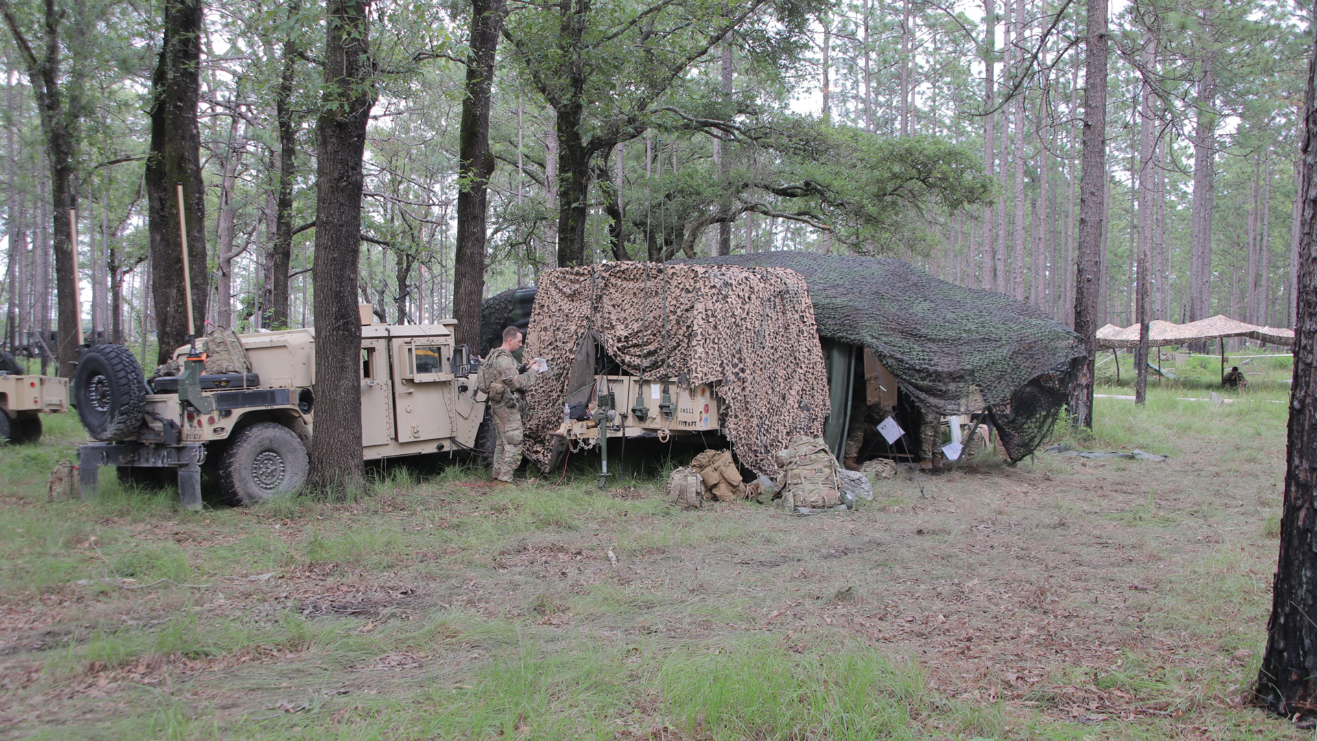 Military field command post in wooded area with tan HMMWV (Humvee) vehicle and camouflage netting shelter. Soldier in combat uniform standing between vehicle and tent structure covered with desert camouflage net and green tarp. Pine forest setting typical of military training area or field exercise location.