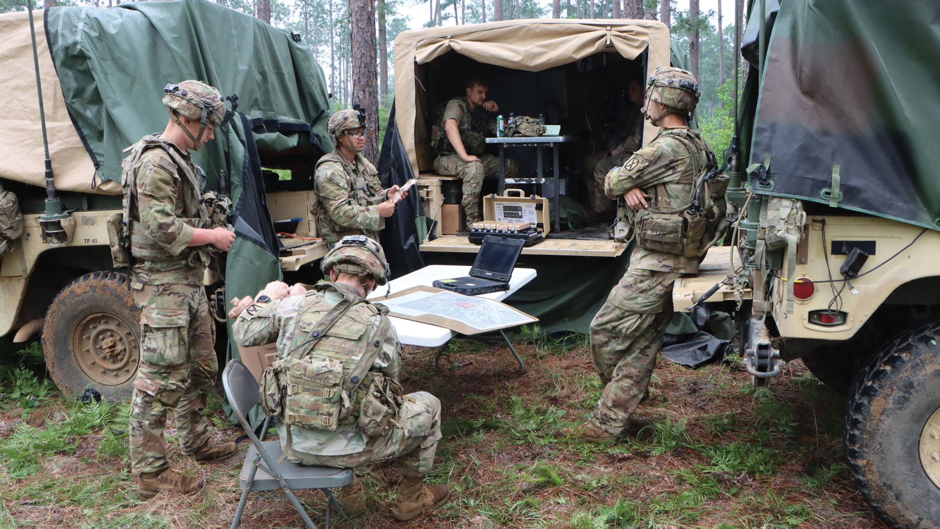 Military communications operations in field environment showing five soldiers in multicam combat uniforms working at mobile command vehicle. Soldier seated at folding table with laptop computer and documents, while others operate equipment from vehicle's open rear compartment containing communications gear. Multiple military vehicles with canvas covers visible in wooded training area.