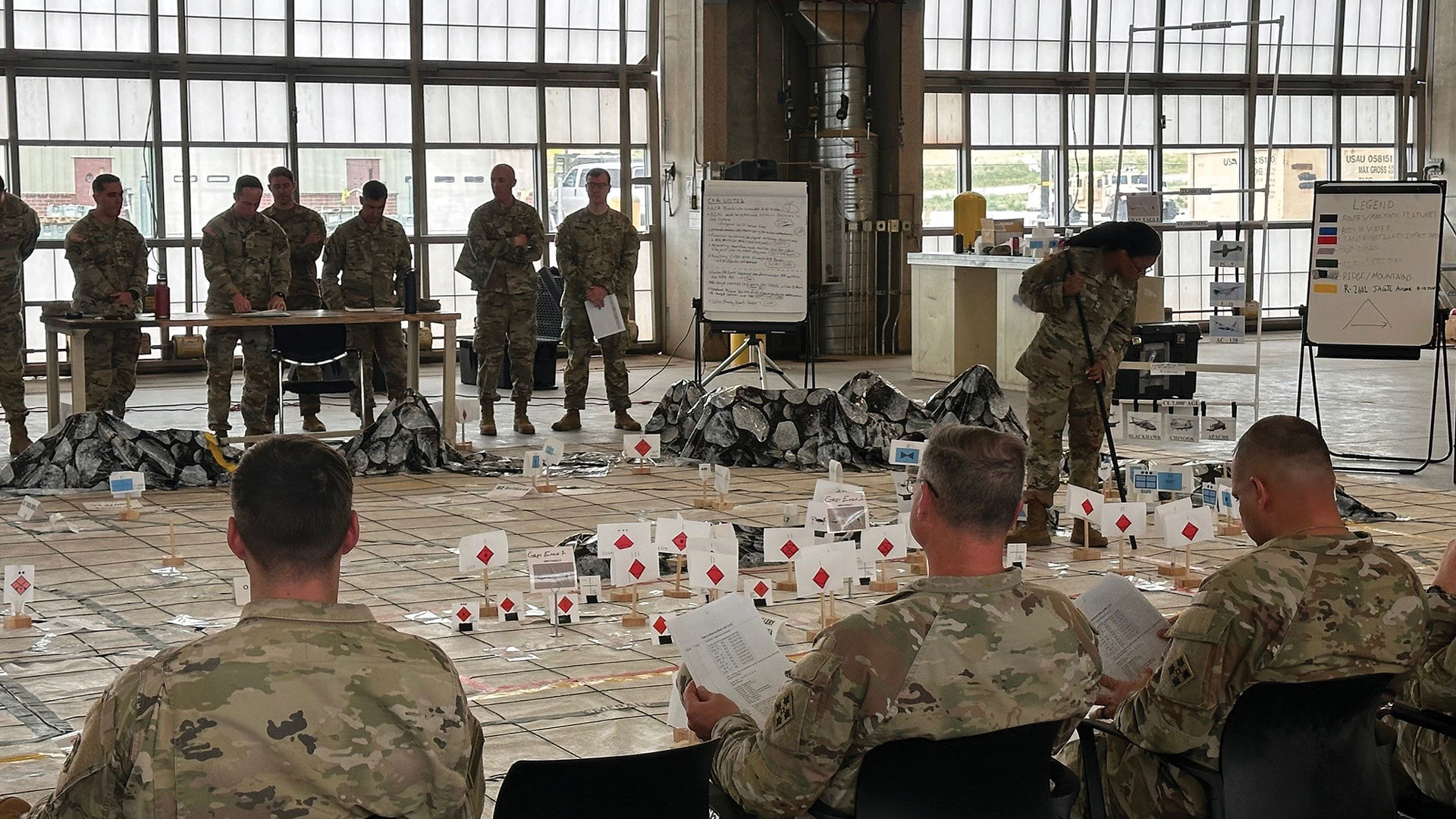 Military training exercise in hangar showing soldiers conducting tabletop wargaming or tactical simulation. Multiple soldiers in combat uniforms positioned around large floor map with white building models marked with red diamond symbols. Briefing board and equipment bags visible, with additional soldiers observing from tables. Industrial hangar setting with large windows providing natural light.