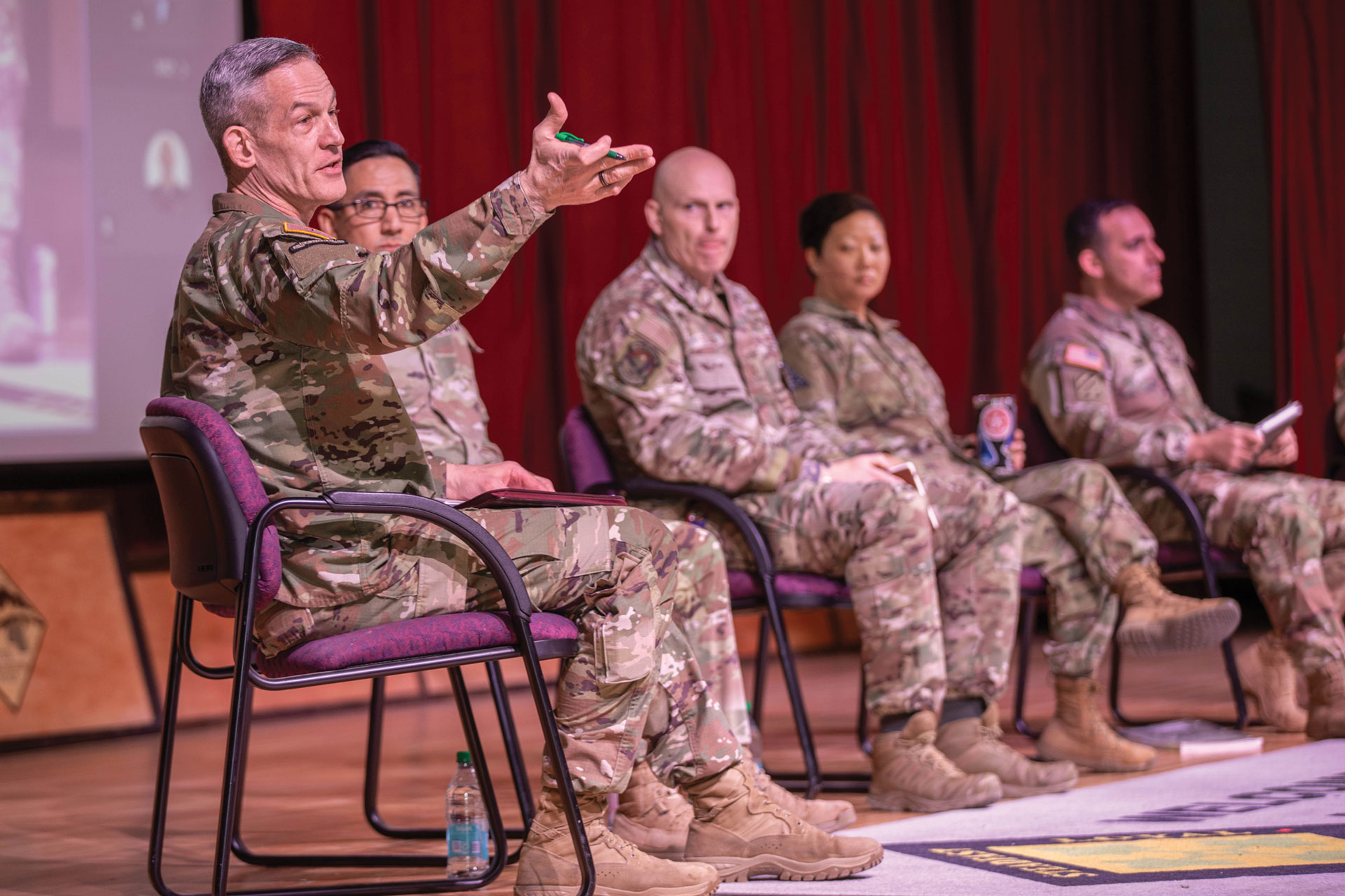 Military panel discussion with five senior officers in combat uniforms seated on stage. Lead officer gesturing while speaking, with four other officers seated in purple chairs against red curtain backdrop. Presentation screen visible, water bottles on floor, suggesting military conference or briefing setting.