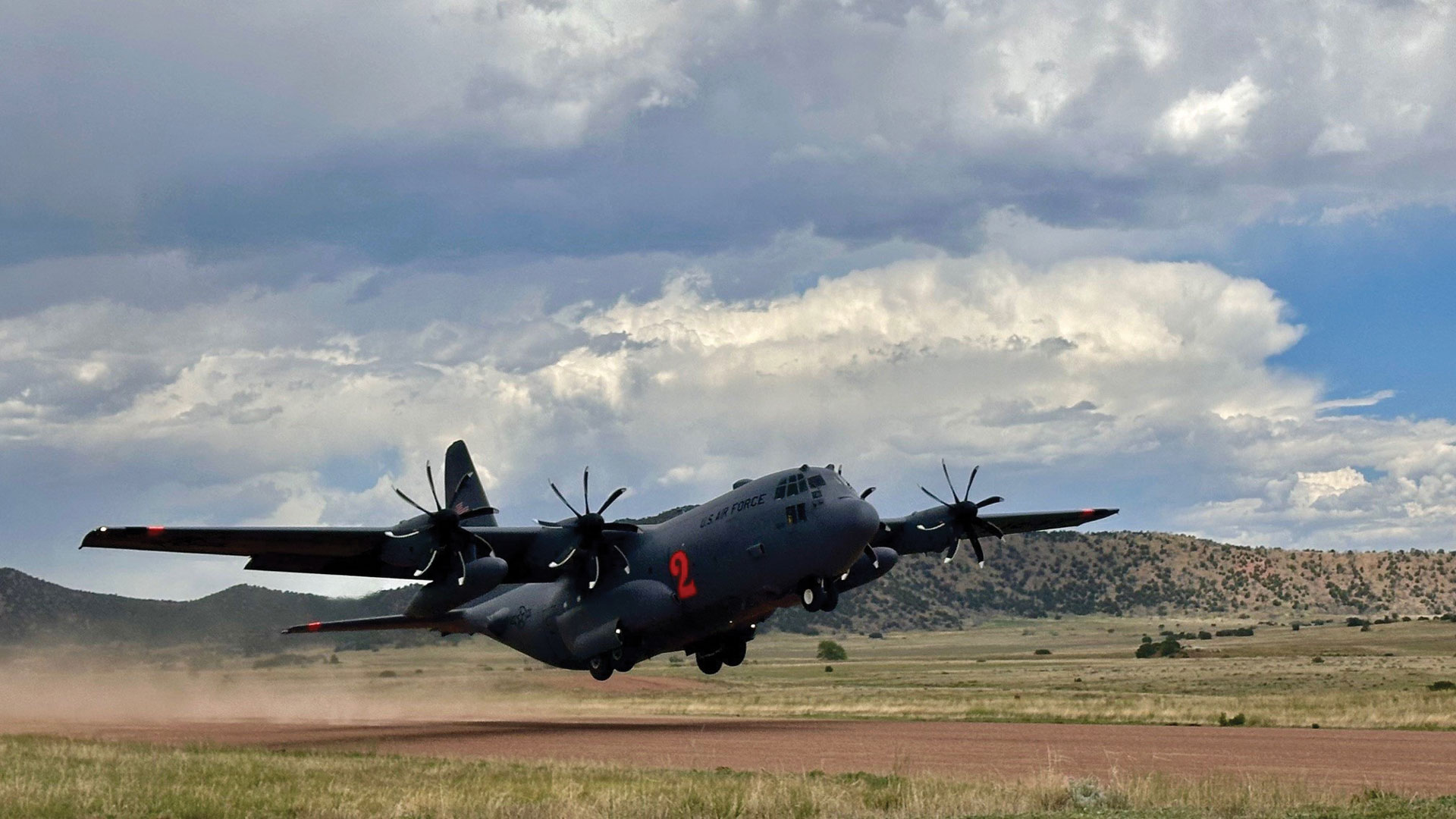 C-130 Hercules aircraft with number 2 marking taking off or landing on dirt airstrip. Four-engine turboprop military transport aircraft against dramatic cloudy sky with desert hills in background. Dust visible from unpaved runway operations.