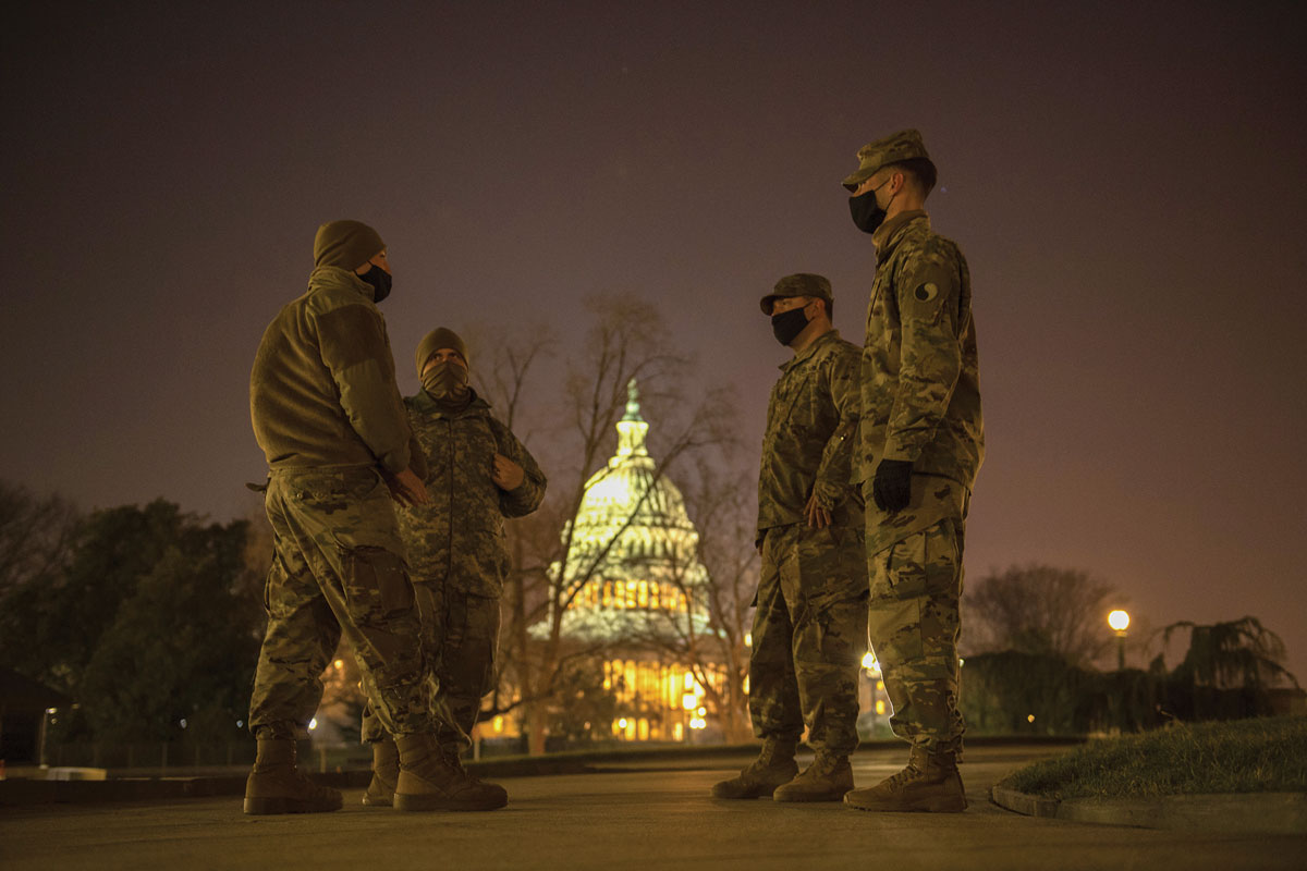 
Four military personnel in fatigues and masks stand conversing at night, with the illuminated Capitol Building in the background.