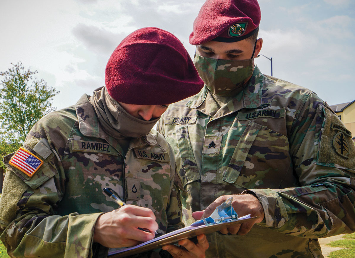 Two U.S. Army soldiers, one writing on a clipboard, are focused on their task. Both are wearing maroon berets, indicating airborne forces, with the background suggesting a daytime outdoor setting.