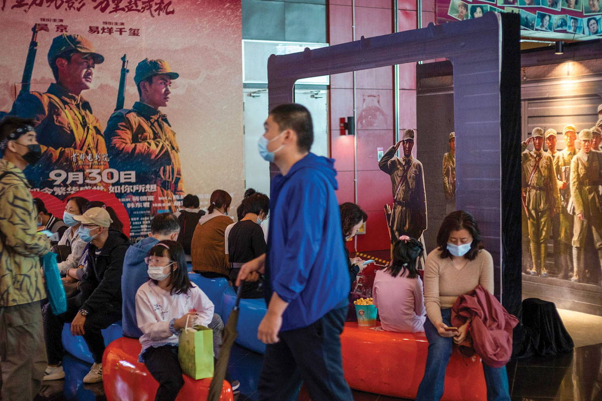 The image shows people in a public setting, possibly a cinema, with a large movie poster in the background featuring military figures and Chinese text, indicating a patriotic theme. Individuals are wearing masks, suggesting the photo was taken during a time when health precautions were common.