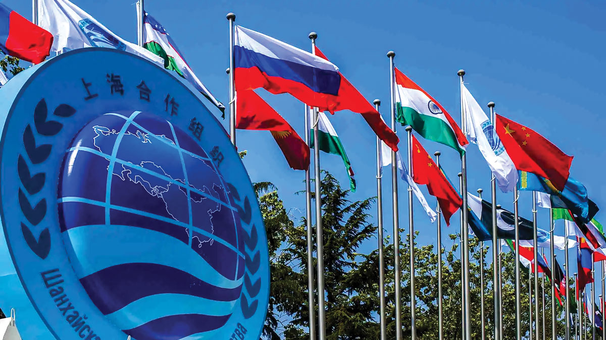 The image shows a collection of international flags flapping in the wind, with a prominent emblem in the foreground displaying Chinese characters, suggesting a global or multinational context, likely an international organization or event in a Chinese-speaking region.