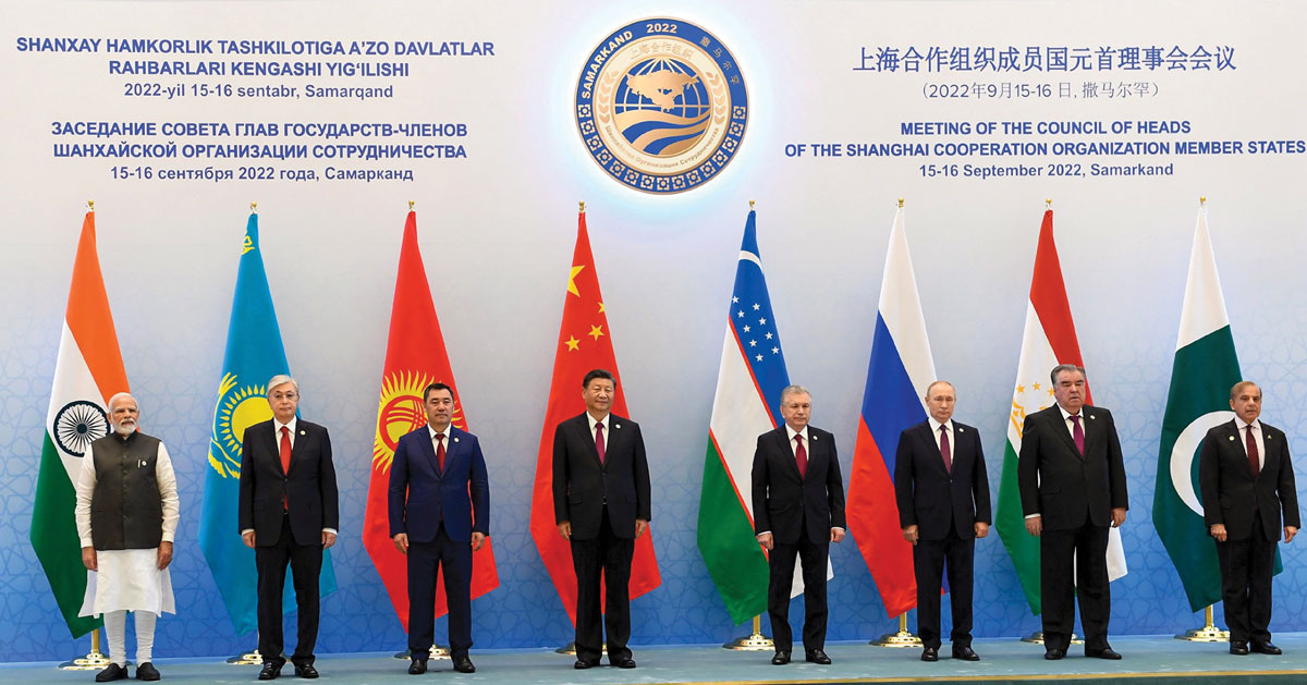 A group of leaders from different nations stand in front of their respective flags, with a banner overhead announcing a meeting of the Shanghai Cooperation Organization (SCO) member states, dated 15-16 September 2022 in Samarkand. The logo of the SCO is displayed prominently above them.