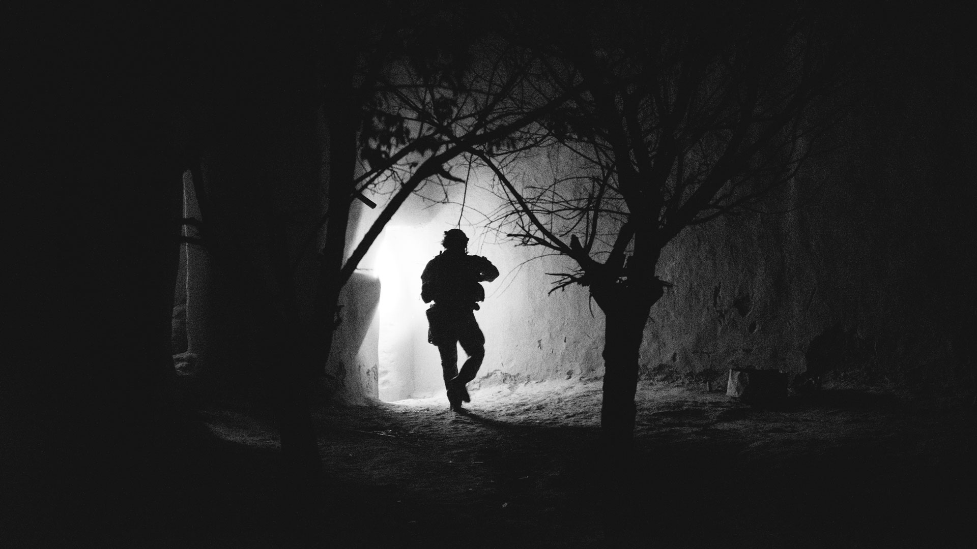 Silhouette of a soldier entering a doorway during a nighttime operation, illuminated by a bright light in a dark, forested environment.