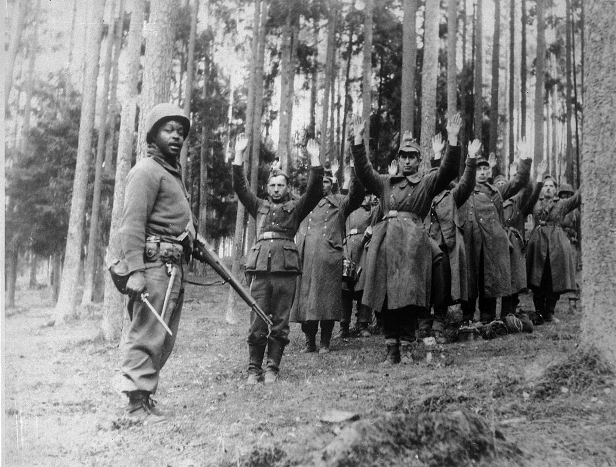 A soldier of the 12th Armored Division stands guard over a group of Nazi prisoners circa April 1945