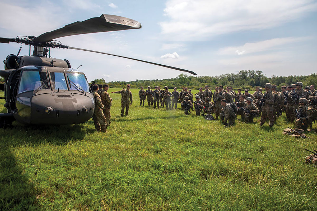 
A group of military personnel in a field with a Black Hawk helicopter. Some are kneeling, others standing; one is pointing, possibly giving instructions. The setting suggests a briefing or debriefing of a mission.