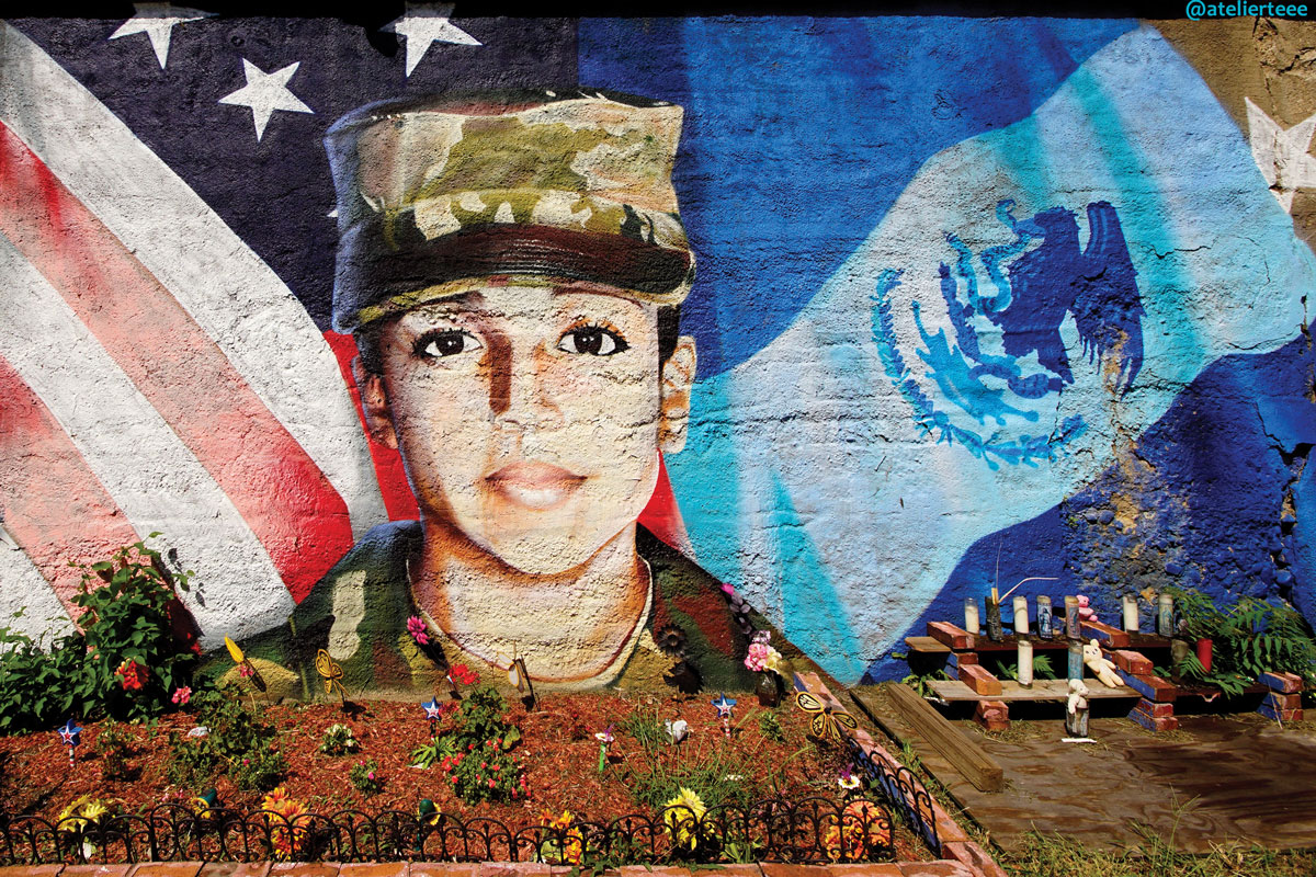 Mural of a soldier's face superimposed on flags, with a small memorial and flowers in the foreground.