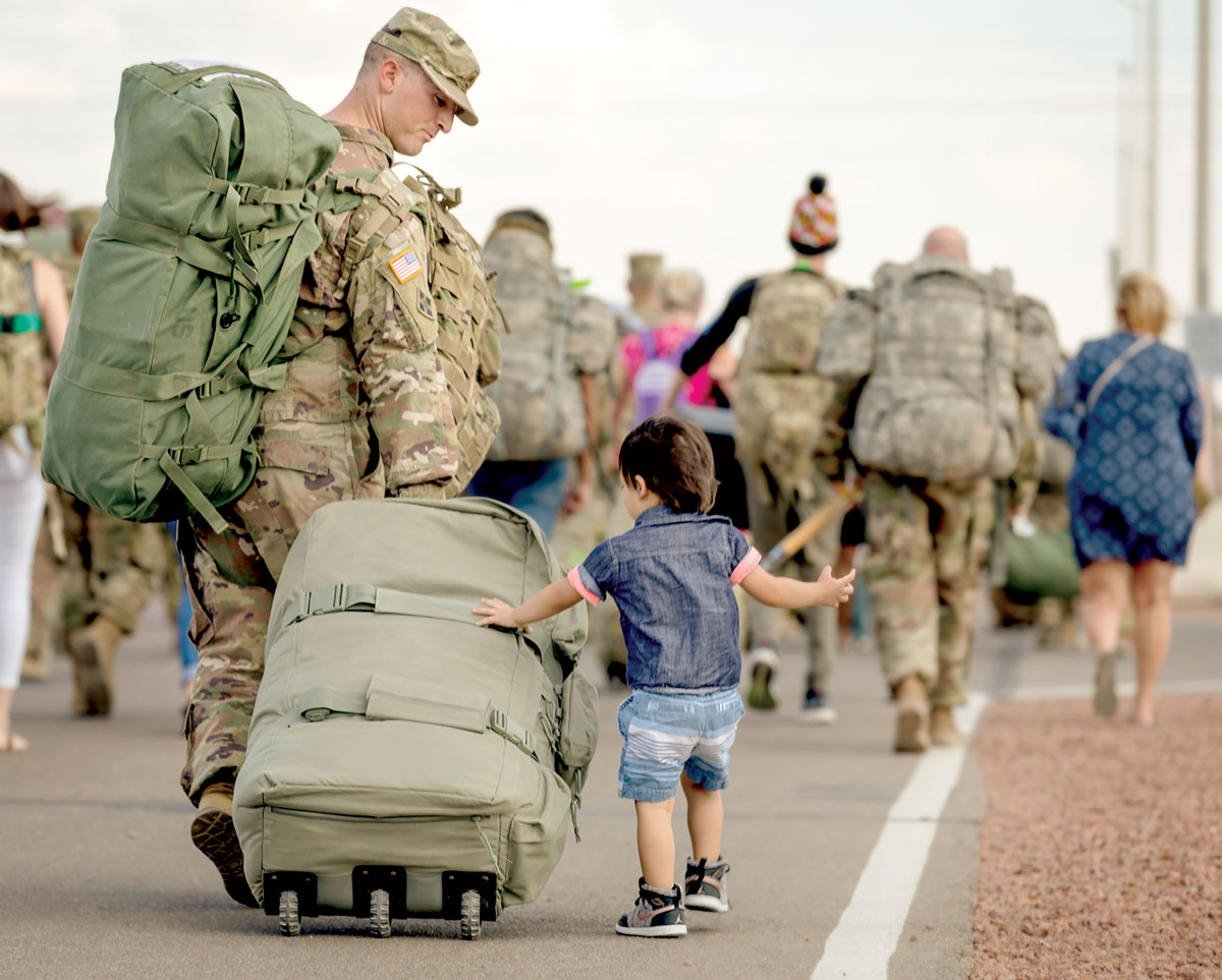 A young child walks beside a uniformed soldier, reaching out to touch a large, wheeled duffle bag. The soldier, laden with gear, glances down at the child. Other military personnel with heavy packs walk ahead.
