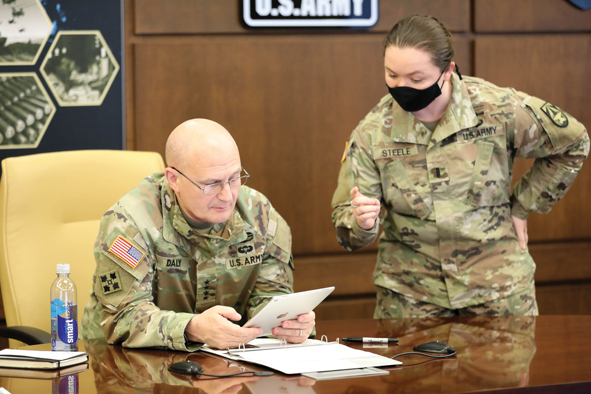 Two U.S. Army soldiers in a meeting room. One, seated, is focused on a tablet, while the other, standing with a mask on, appears to be speaking. Documents and a water bottle are on the table.
