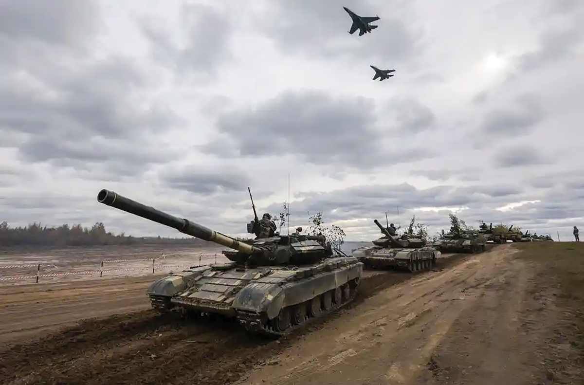 Line of armored tanks positioned on a dirt road with fighter jets flying overhead against a cloudy sky.