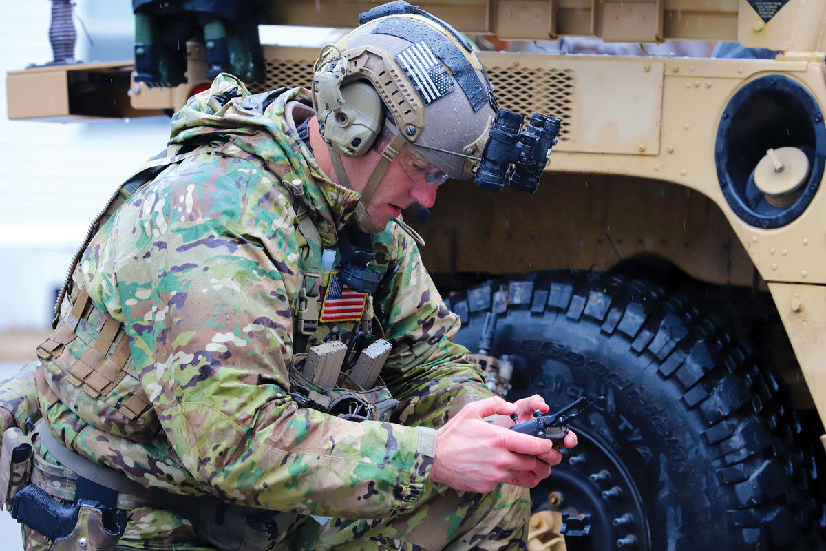 A soldier in camouflage gear focuses on a handheld device, with a military vehicle in the background.