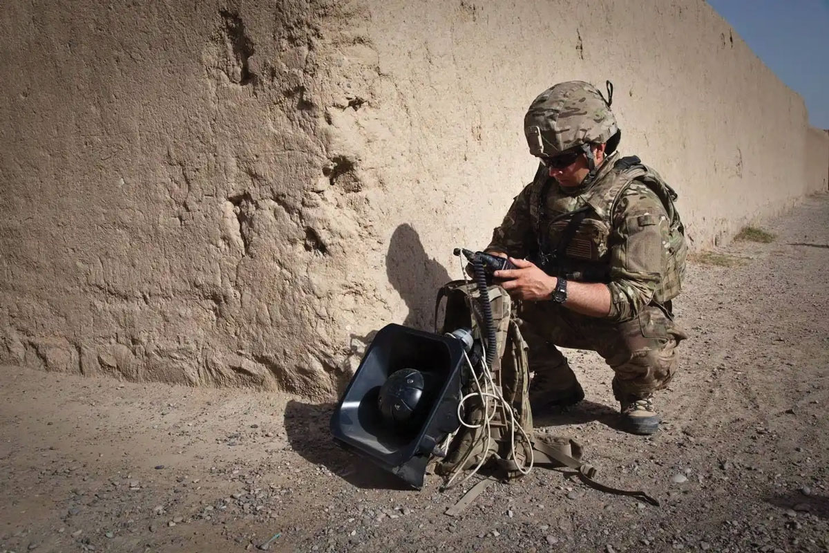 A soldier crouched down, inspecting military equipment beside an adobe wall