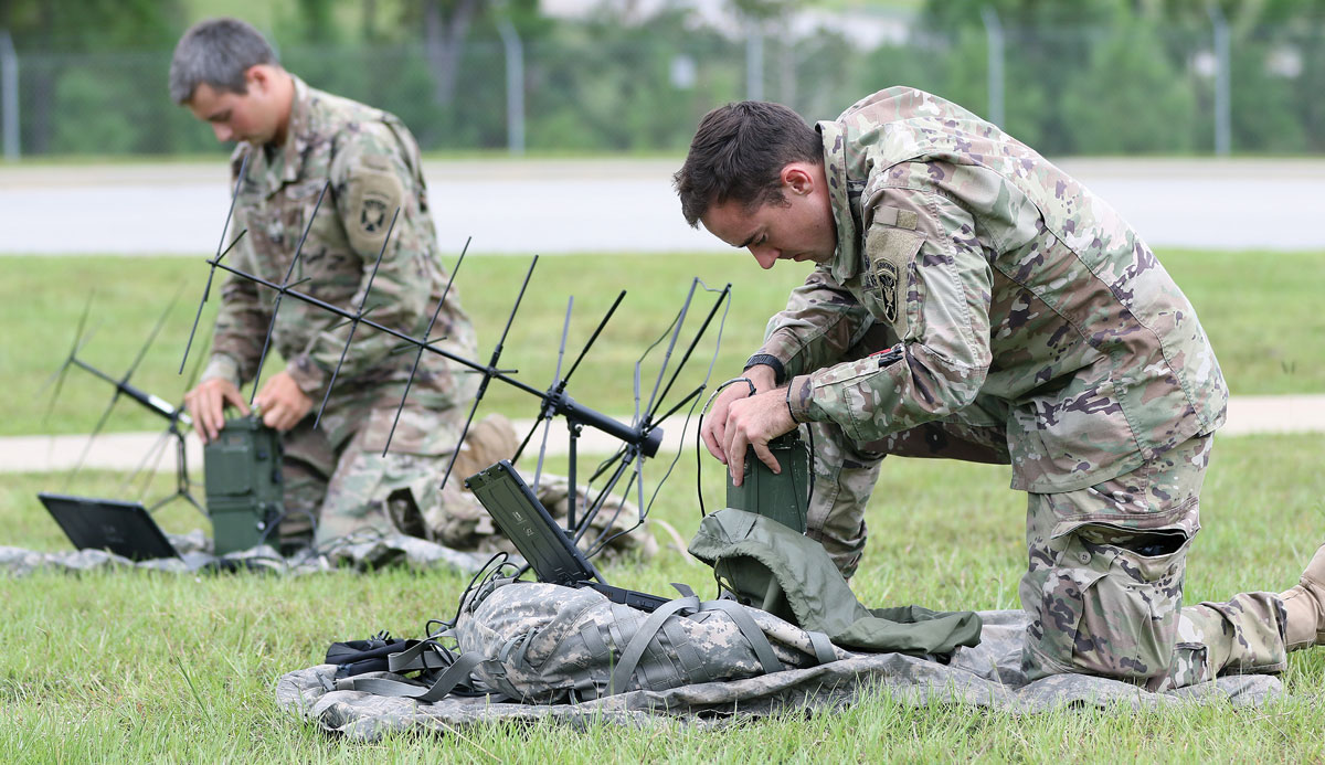 Two soldiers in camouflage setting up communication equipment on a grassy field.