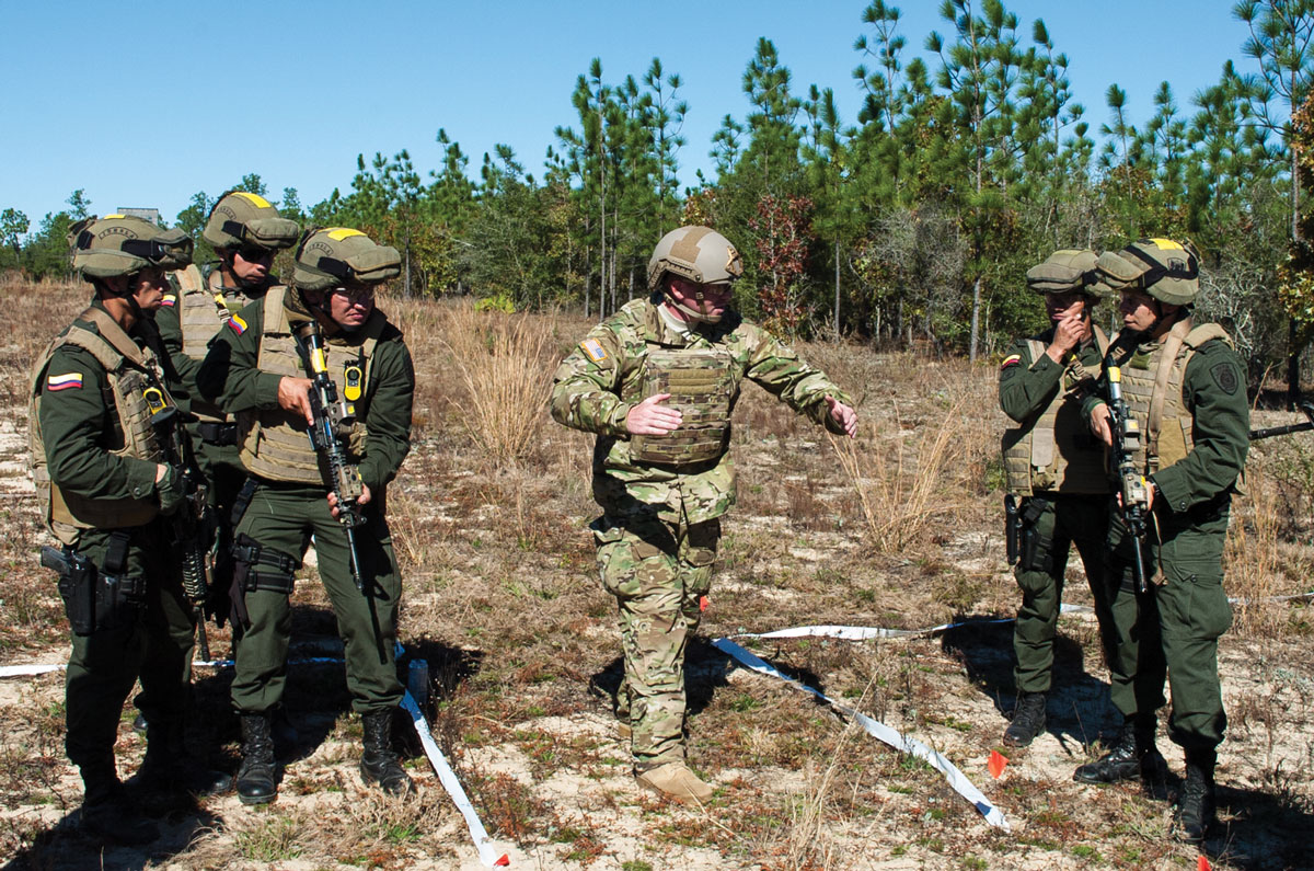 A group of soldiers in combat gear engaged in an outdoor briefing with one gesturing instructions.