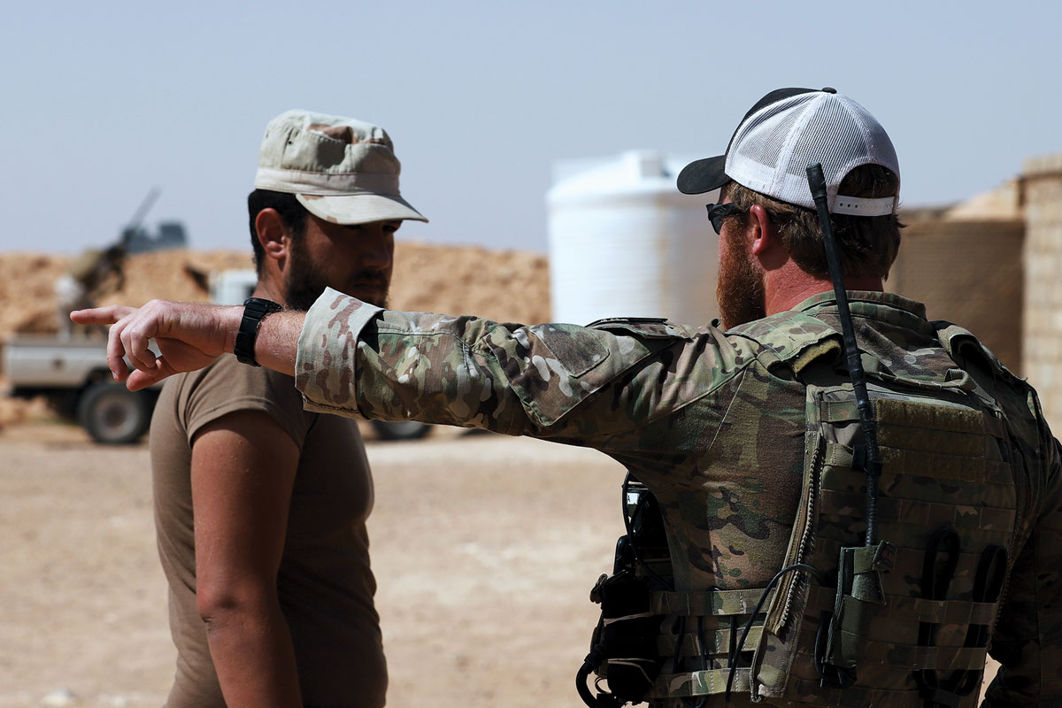 An Army “Green Beret” pointing directions to a Maghaweir al-Thowra (MaT) soldier in a desert setting, with military vehicles in the background.