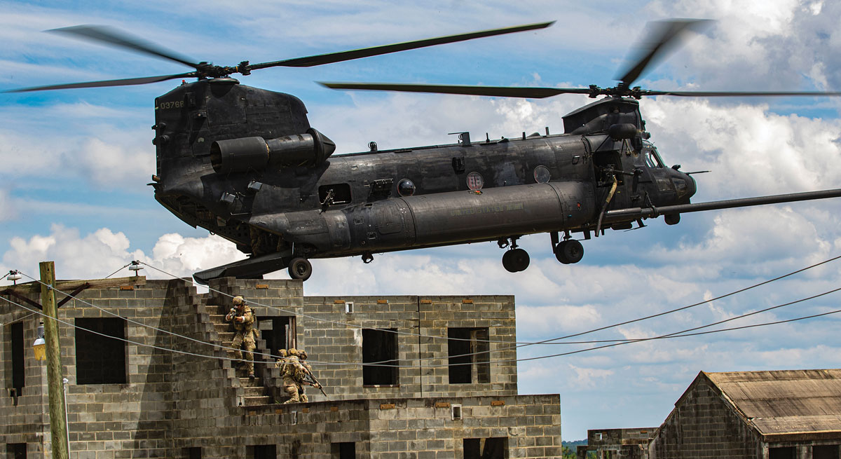 A military helicopter hovers above urban structures with soldiers in the scene.