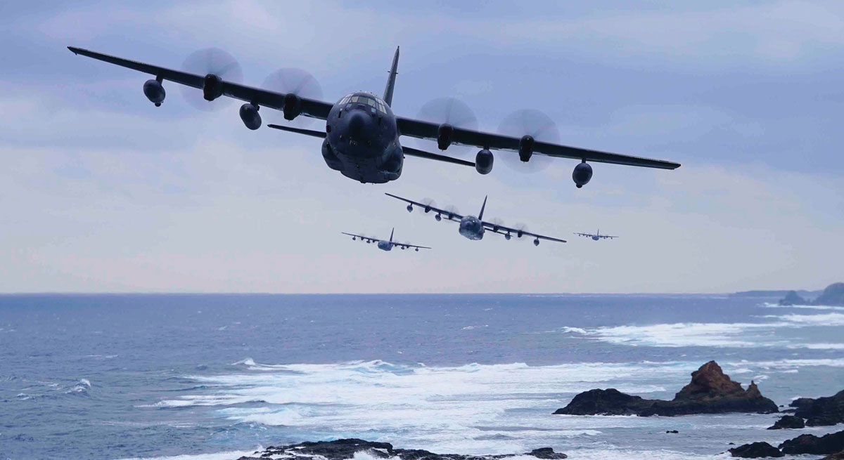 Military transport aircraft flying in formation over a coastal landscape with rocky shores and ocean waves.