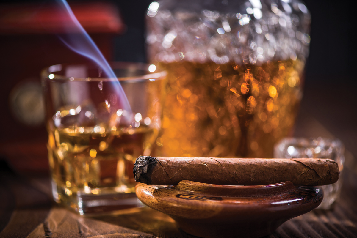 a close-up of a lit cigar resting on an ashtray with a glass of whiskey in the background