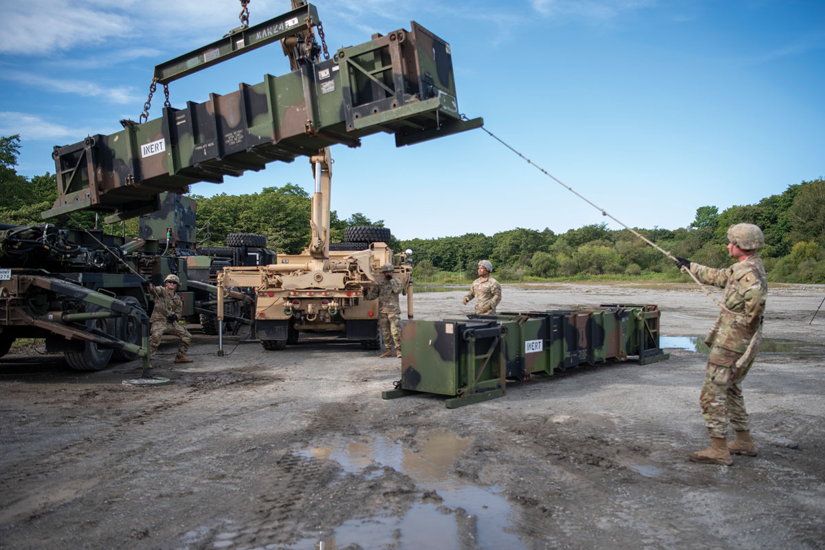 soldiers using a crane to load a large green container onto a military transporter in a field, under a clear sky.